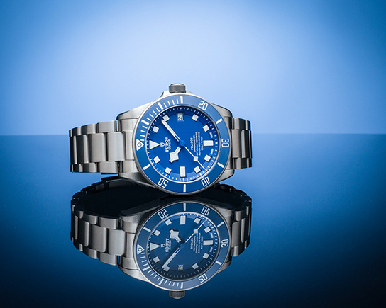 Luxury Tudor dive watch photographed on reflective blue surface with precision studio lighting for high-end commercial product photography.